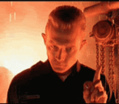 A Man In A Black Shirt Is Giving The Middle Finger In Front Of A Fire . GIF