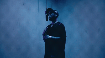 A Man In A Black Shirt Is Holding A Microphone In Front Of A Blue Wall GIF