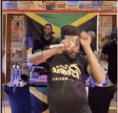 A Man In A Black Shirt Is Holding A Microphone In Front Of A Flag . GIF