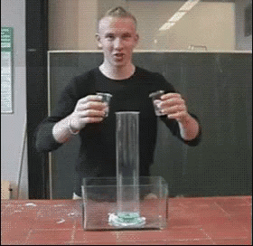 A Man In A Black Shirt Is Holding Two Shot Glasses In Front Of A Glass Container GIF