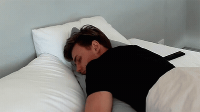 A Man In A Black Shirt Is Laying On A Bed GIF