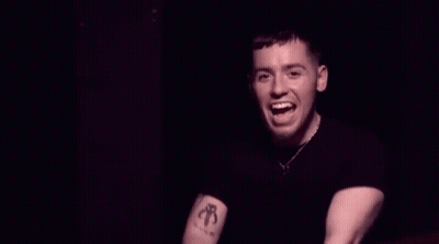 A Man In A Black Shirt Is Pointing At The Camera And Smiling In A Dark Room . GIF