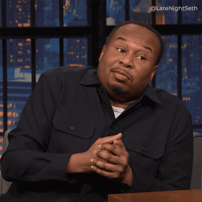 A Man In A Black Shirt Is Sitting At A Table With His Hands Outstretched In Front Of A Screen That Says Latenightseth GIF