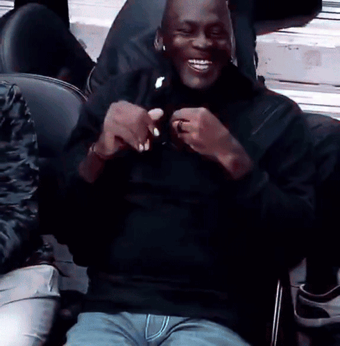 A Man In A Black Shirt Is Sitting In A Chair And Smiling . GIF