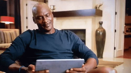 A Man In A Black Shirt Is Sitting In A Chair Holding A Tablet GIF