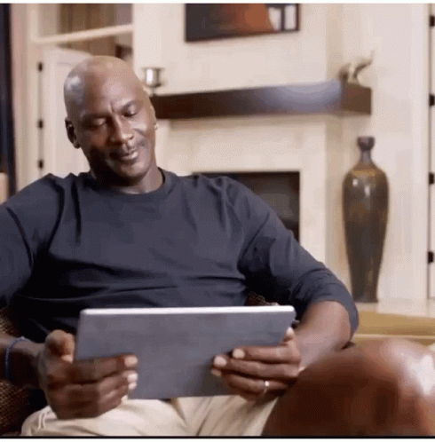 A Man In A Black Shirt Is Sitting In A Chair Looking At A Tablet GIF