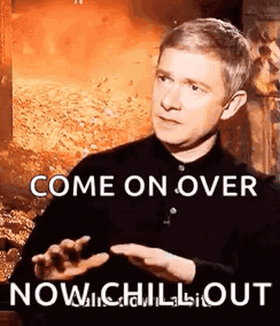 A Man In A Black Shirt Is Sitting In Front Of A Fireplace And Talking To Someone While Holding His Hand Out . GIF