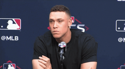 A Man In A Black Shirt Is Sitting In Front Of A Mlb Sign GIF