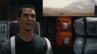 A Man In A Black Shirt Is Sitting In Front Of A Screen With A Picture Of A Desert On It GIF