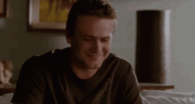 A Man In A Black Shirt Is Sitting On A Couch And Smiling . GIF