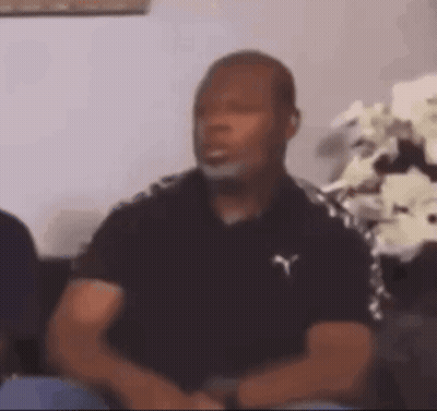 A Man In A Black Shirt Is Sitting On A Couch In Front Of A Vase Of Flowers . GIF