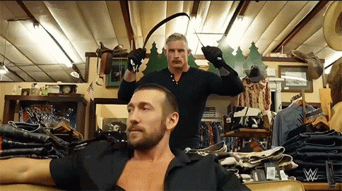 A Man In A Black Shirt Is Sitting On A Couch While Another Man Holds A Whip In A Store GIF
