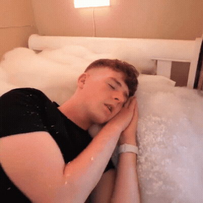 A Man In A Black Shirt Is Sleeping In A Bed Covered In Foam GIF