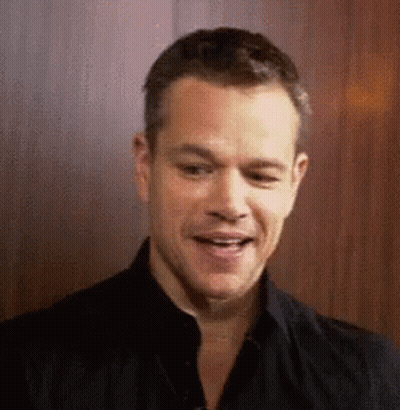 A Man In A Black Shirt Is Smiling And Looking At The Camera While Standing In Front Of A Wooden Wall . GIF