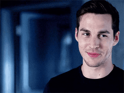 A Man In A Black Shirt Is Smiling And Looking At The Camera With A Blue Background . GIF