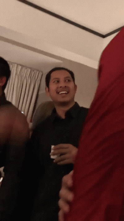 A Man In A Black Shirt Is Smiling In A Room With Other People GIF