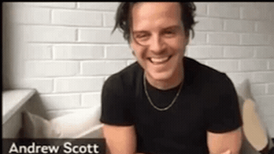 A Man In A Black Shirt Is Smiling In Front Of A Brick Wall . GIF