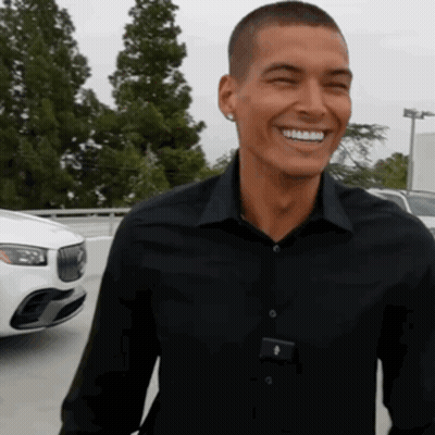 A Man In A Black Shirt Is Smiling In Front Of A White Car In A Parking Lot GIF