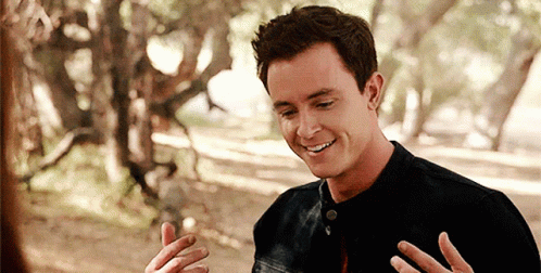 A Man In A Black Shirt Is Smiling With His Hands Outstretched In Front Of Trees GIF