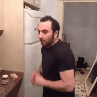 A Man In A Black Shirt Is Standing In A Kitchen Next To A Refrigerator . GIF