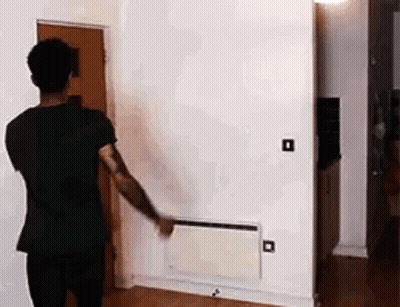 A Man In A Black Shirt Is Standing In A Living Room Looking At A Wall Heater . GIF