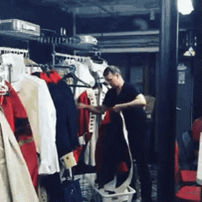 A Man In A Black Shirt Is Standing In A Room With Clothes Hanging On Racks GIF