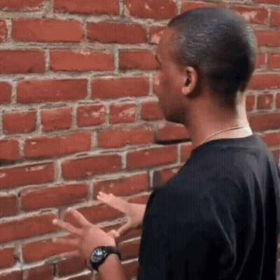 A Man In A Black Shirt Is Standing In Front Of A Brick Wall With His Hands Outstretched . GIF