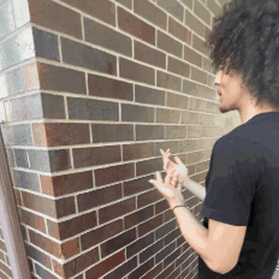 A Man In A Black Shirt Is Standing In Front Of A Brown Brick Wall GIF