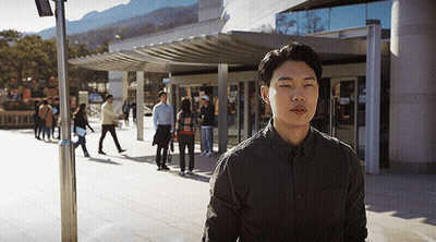 A Man In A Black Shirt Is Standing In Front Of A Building With People Walking In The Background GIF