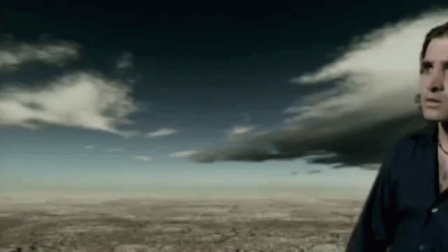 A Man In A Black Shirt Is Standing In Front Of A Cloudy Sky GIF