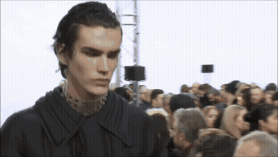 A Man In A Black Shirt Is Standing In Front Of A Crowd Of People GIF