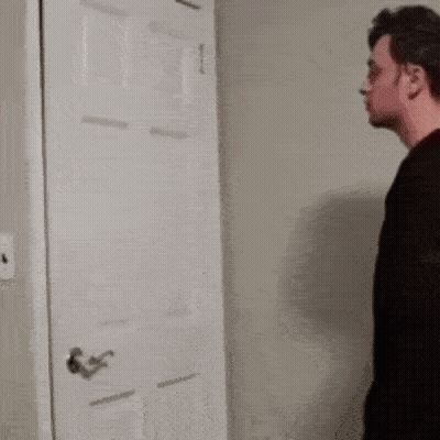 A Man In A Black Shirt Is Standing In Front Of A Door . GIF