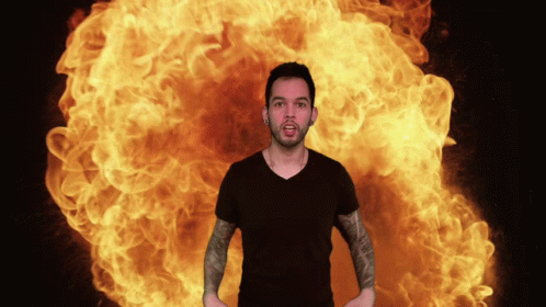 A Man In A Black Shirt Is Standing In Front Of A Large Explosion Of Flames GIF