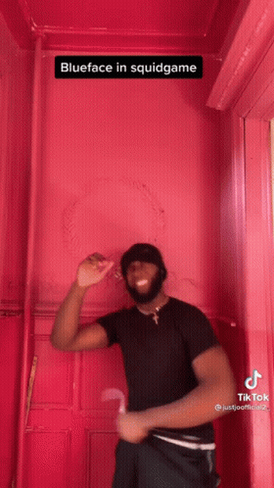 A Man In A Black Shirt Is Standing In Front Of A Pink Wall With The Caption Blueface In Squidgame . GIF