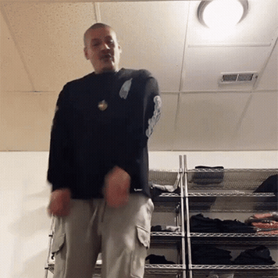 A Man In A Black Shirt Is Standing In Front Of A Rack Of Clothes GIF