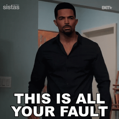 A Man In A Black Shirt Is Standing In Front Of A Sign That Says This Is All Your Fault GIF