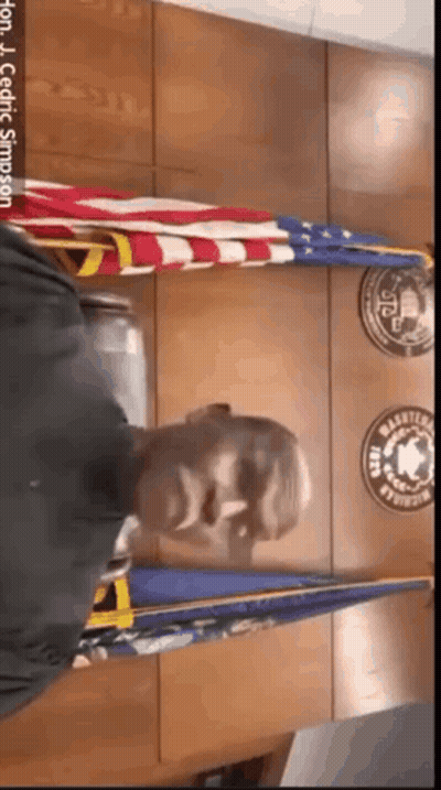 A Man In A Black Shirt Is Standing In Front Of A Wall With Flags And Badges On It .. GIF