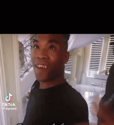 A Man In A Black Shirt Is Standing In Front Of A Window With Blinds . GIF