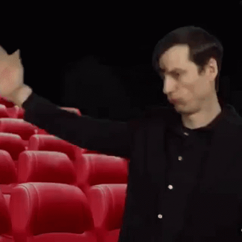 A Man In A Black Shirt Is Standing In Front Of Red Seats In A Theater . GIF
