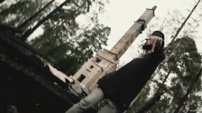 A Man In A Black Shirt Is Standing In The Woods In Front Of A Statue . GIF