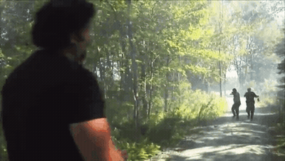 A Man In A Black Shirt Is Standing On A Dirt Road In The Woods Looking At Two Men Walking Down The Road . GIF