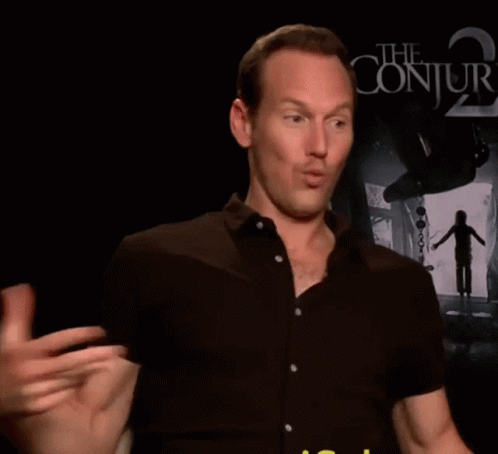 A Man In A Black Shirt Is Talking In Front Of A Poster For The Conjuring 2 GIF