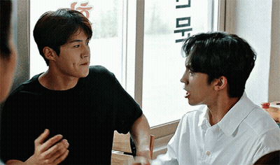 A Man In A Black Shirt Is Talking To Another Man In A White Shirt While Sitting In Front Of A Window . GIF