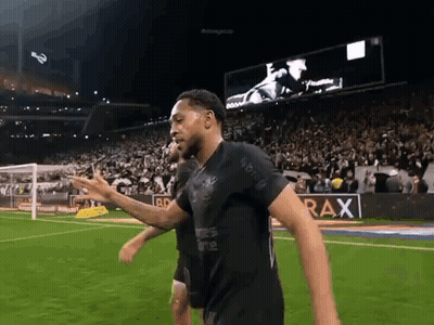 A Man In A Black Shirt Is Walking On A Soccer Field GIF