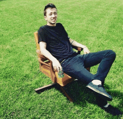 A Man In A Black Shirt Sits In A Wooden Chair In The Grass GIF