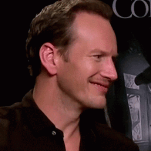 A Man In A Black Shirt Smiles In Front Of A Sign That Says Co GIF