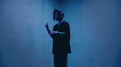 A Man In A Black Shirt Stands In Front Of A Blue Wall Holding A Microphone GIF