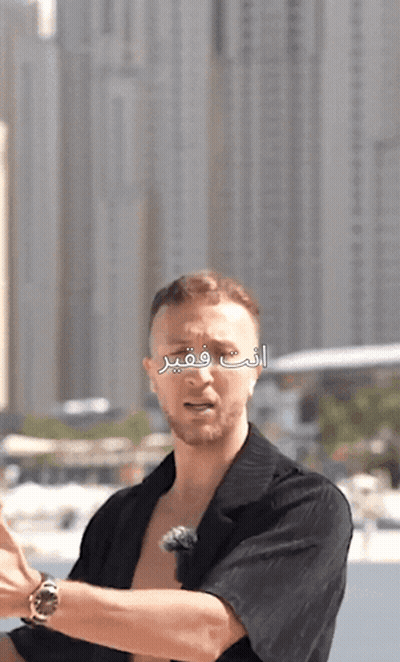 A Man In A Black Shirt Stands In Front Of A City Skyline With Arabic Writing On It GIF