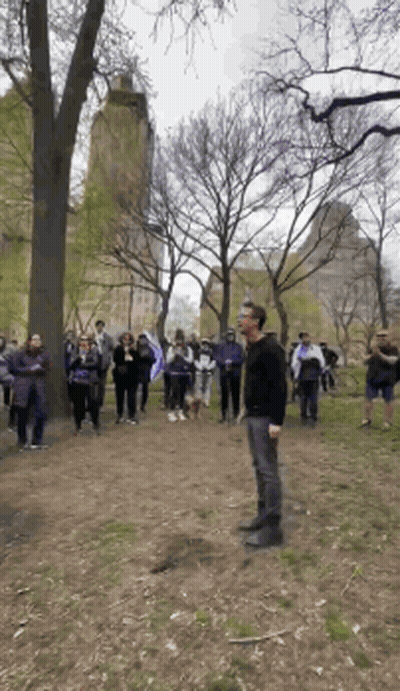A Man In A Black Shirt Stands In Front Of A Crowd In A Park GIF