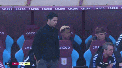 A Man In A Black Shirt Stands In Front Of A Row Of Purple And Blue Seats That Say Cazoo GIF